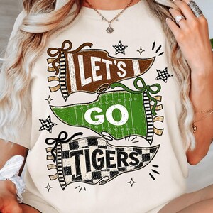 May include: A cream-colored t-shirt featuring a graphic design of three pennants. The top brown pennant says LET'S, the middle green football field pennant says GO, and the bottom checkered pennant says TIGERS. Small black stars and sparkles surround the text.