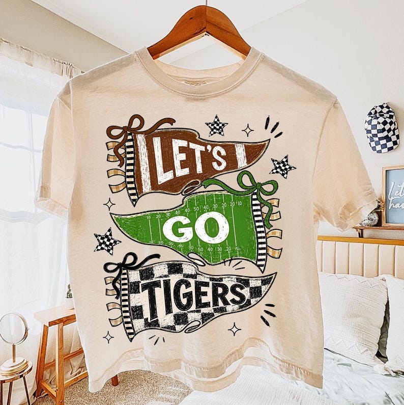 May include: A cream-colored short-sleeve t-shirt featuring a graphic print of three pennants stacked vertically. The top brown pennant reads "LET'S", the middle green football field pennant reads "GO", and the bottom checkered pennant reads "TIGERS".