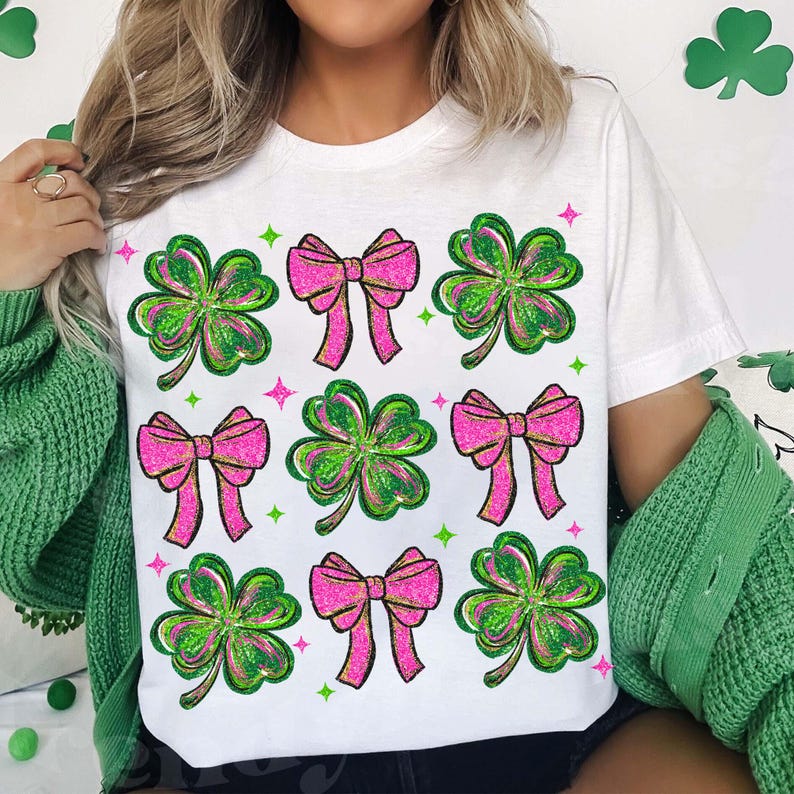 May include: White t-shirt featuring a pattern of pink bows and green shamrocks. The bows have a glittery texture, and the shamrocks are outlined in green with a glittery effect. Green and pink star accents are scattered around the design.