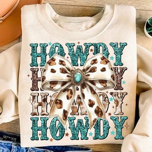 May include: A cream-coloured sweatshirt with a graphic design of the word "Howdy" repeated four times in a turquoise, brown, and white colour scheme. The word "Howdy" is surrounded by a large brown and white leopard print bow with a turquoise gemstone in the centre.
