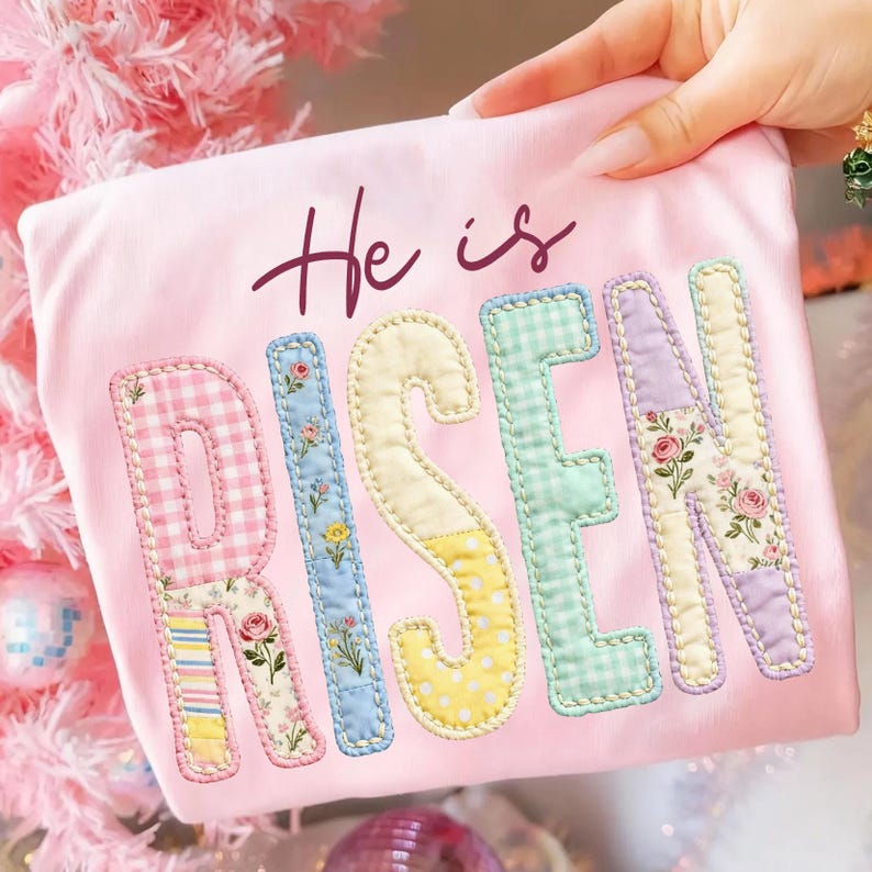 May include: A light pink t-shirt with the words "He is RISEN" in a patchwork design. The letters are in various pastel colors and patterns, including gingham, floral, and polka dots. The text is in a handwritten style.