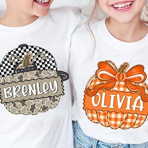 Op de afbeelding: Twee witte t-shirts met pompoenafbeeldingen. Het linker t-shirt toont een camouflagepompoen met een zwart-wit geblokte honkbalpet en de naam Brenley. Het rechter t-shirt toont een oranje geruite pompoen met een strik en de naam Olivia.