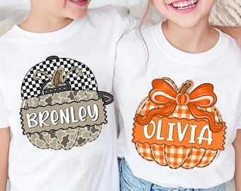 May include: Two white t-shirts featuring pumpkin graphics. The left shirt displays a camouflage pumpkin wearing a black and white checkered baseball cap with the name Brenley. The right shirt shows an orange plaid pumpkin with a bow and the name Olivia.