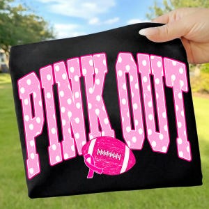 May include: A black t-shirt featuring the text PINK OUT in large, pink, block-style letters with a white polka dot pattern. Below the text is a pink American football graphic with a white ribbon symbol integrated into the design.