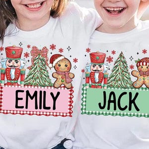 Puede incluir: Camisetas blancas con diseños navideños. Una camiseta presenta "EMILY" en una pancarta rosa con un cascanueces, un árbol de Navidad y un hombre de jengibre. La otra camiseta tiene "JACK" en una pancarta verde con diseños festivos similares.