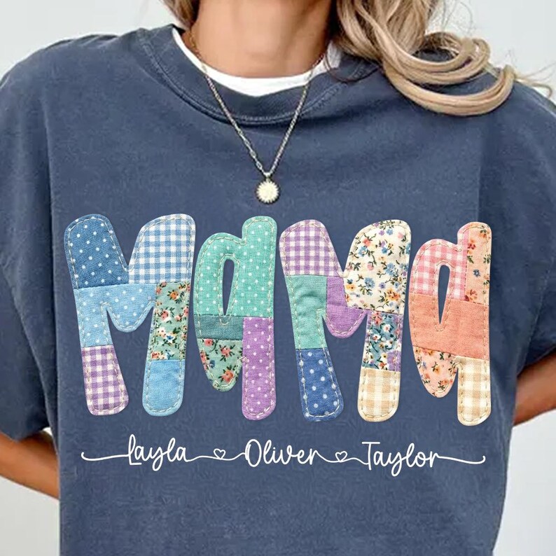 May include: A navy blue sweatshirt features the word "Mama" in a patchwork design with various patterns and colors. Below the word are the names "Layla", "Oliver", and "Taylor" in a cursive font.