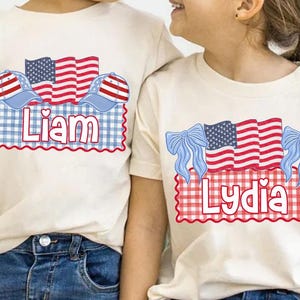 Puede incluir: Dos camisetas color crema con dise&ntilde;os patri&oacute;ticos. Una camiseta presenta el nombre "Liam" con una bandera roja, blanca y azul y un dise&ntilde;o de gorra de b&eacute;isbol. La otra camiseta tiene el nombre "Lydia" con un dise&ntilde;o de bandera y lazo similar.