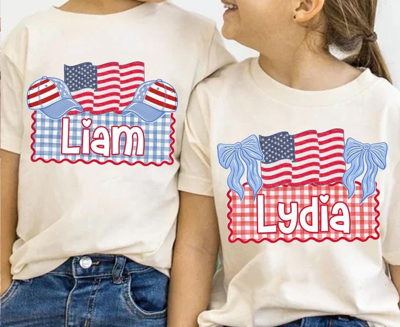 Puede incluir: Dos camisetas color crema con dise&ntilde;os patri&oacute;ticos. Una camiseta presenta el nombre "Liam" con una bandera roja, blanca y azul y un dise&ntilde;o de gorra de b&eacute;isbol. La otra camiseta tiene el nombre "Lydia" con un dise&ntilde;o de bandera y lazo similar.