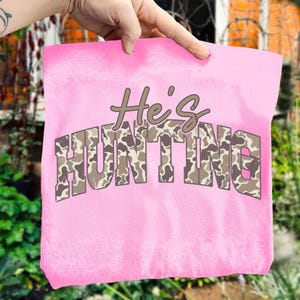 May include: A pink tote bag with the text "He's HUNTING" in brown and camouflage lettering. The bag is held up by a hand, showcasing the design. The background is blurred, with greenery and a brick wall visible.