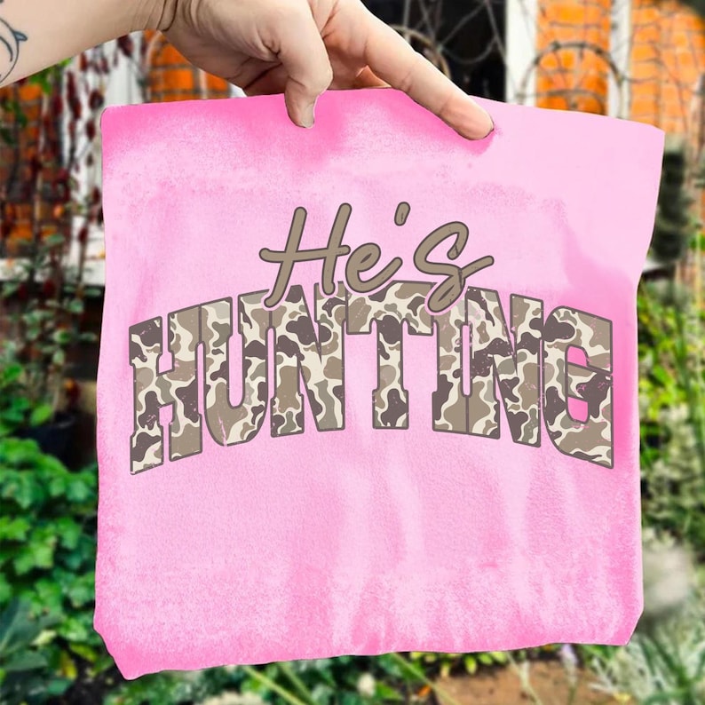 May include: A pink tote bag with the text "He's HUNTING" in brown and camouflage lettering. The bag is held up by a hand, showcasing the design. The background is blurred, with greenery and a brick wall visible.