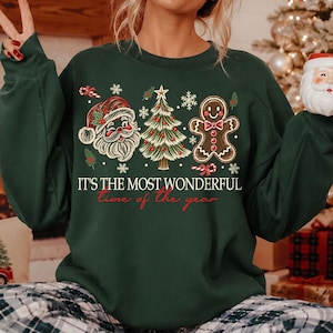 K&ouml;nnte beinhalten: Ein dunkelgr&uuml;nes Langarm-Sweatshirt mit einem Grafikdruck von Weihnachtsmann, einem geschm&uuml;ckten Weihnachtsbaum und einem Lebkuchenmann. Der Text lautet IT'S THE MOST WONDERFUL time of the year in wei&szlig;er und roter Schrift mit Schneeflocken.