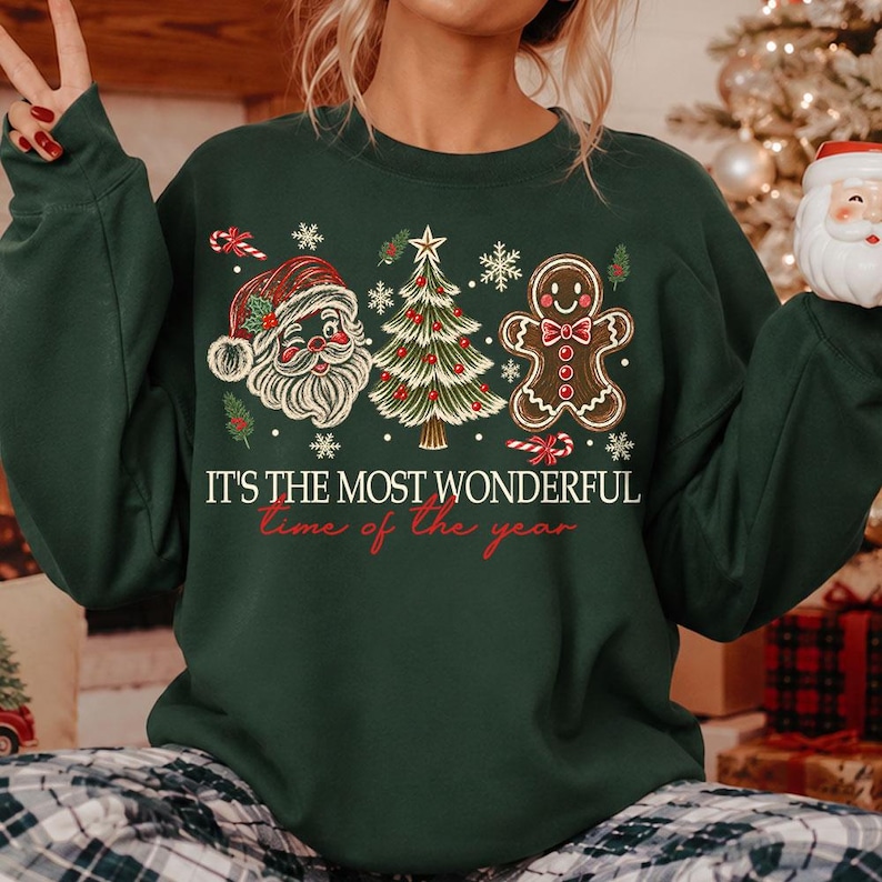 K&ouml;nnte beinhalten: Ein dunkelgr&uuml;nes Langarm-Sweatshirt mit einem Grafikdruck von Weihnachtsmann, einem geschm&uuml;ckten Weihnachtsbaum und einem Lebkuchenmann. Der Text lautet IT'S THE MOST WONDERFUL time of the year in wei&szlig;er und roter Schrift mit Schneeflocken.
