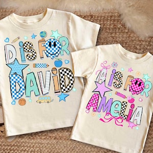 May include: Two cream-colored t-shirts featuring graphic prints. The left shirt displays the text D IS FOR DAVID with a blue checkered smiley face, stars, pencils, and a bus. The right shirt shows A IS FOR AMELIA with a pink checkered smiley face, stars, and bows.