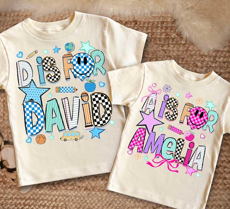 May include: Two cream-colored t-shirts featuring graphic prints. The left shirt displays the text D IS FOR DAVID with a blue checkered smiley face, stars, pencils, and a bus. The right shirt shows A IS FOR AMELIA with a pink checkered smiley face, stars, and bows.