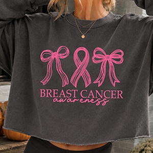 May include: A charcoal gray cropped sweatshirt featuring a pink graphic of two bows flanking a central awareness ribbon. Below the graphic, the text reads BREAST CANCER awareness in a mix of block and script fonts. The garment has a crew neckline.