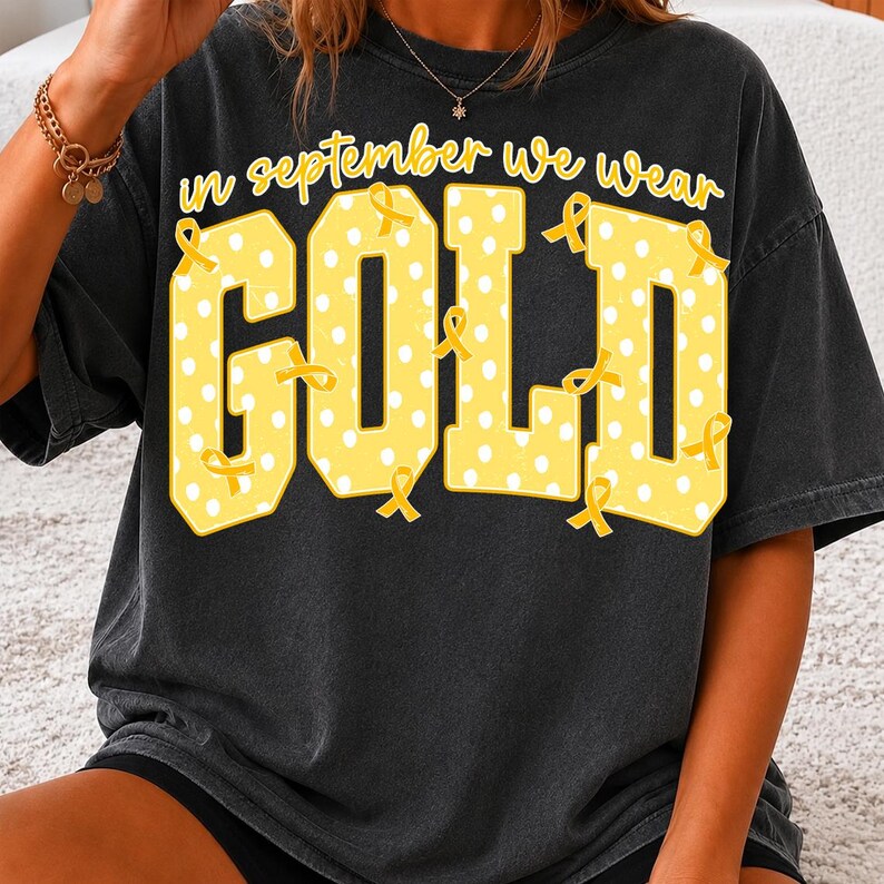 May include: A charcoal gray short-sleeve t-shirt featuring the yellow text "in september we wear GOLD" in a collegiate font. The word "GOLD" is filled with white polka dots and decorated with multiple yellow awareness ribbons throughout.