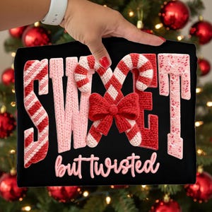 May include: Black t-shirt with the words "SWEET" and "but twisted" in a pink and red knitted design. The design includes candy canes and a bow. The t-shirt is held up in front of a Christmas tree.