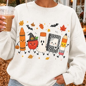 May include: A cream-colored crewneck sweatshirt featuring a graphic print of five anthropomorphic school supplies dressed for Halloween: an orange crayon, an apple with a witch hat, a ghost, a black notebook with a spiderweb, and a pencil.