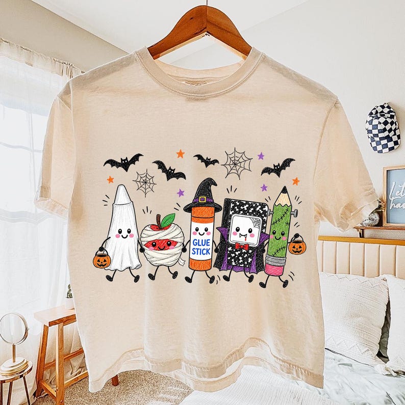 Pode incluir: Uma t-shirt bege de manga curta com uma impress&atilde;o gr&aacute;fica de cinco personagens de desenhos animados vestidos para o Halloween: um fantasma, uma ma&ccedil;&atilde; m&uacute;mia, uma cola em bast&atilde;o com chap&eacute;u de bruxa, um caderno vampiro e um l&aacute;pis.