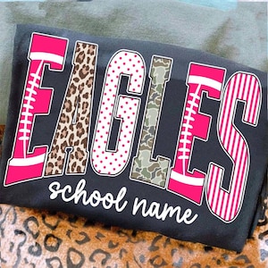 May include: A dark gray t-shirt featuring the word EAGLES in large, multi-patterned letters. The E and S are pink with white football laces, the A is leopard print, the G is white with pink dots, the L is camouflage, and the text school name is below.
