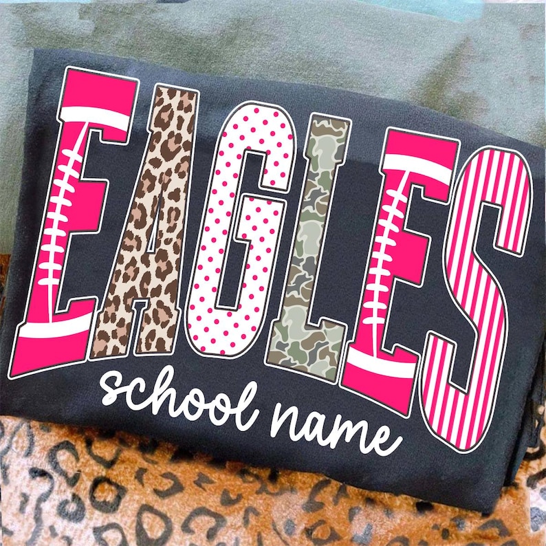 May include: A dark gray t-shirt featuring the word EAGLES in large, multi-patterned letters. The E and S are pink with white football laces, the A is leopard print, the G is white with pink dots, the L is camouflage, and the text school name is below.
