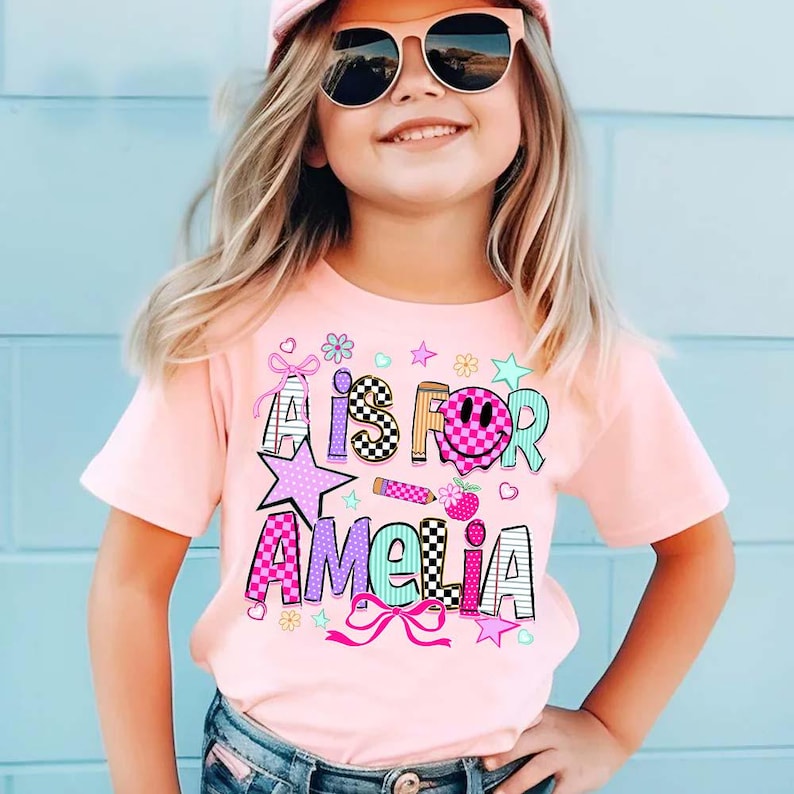 May include: A light pink short-sleeved t-shirt featuring a graphic print with the text A IS FOR AMELIA. The design includes colorful patterns, a checkered smiley face, a pencil, stars, flowers, and bows in shades of pink, blue, and black.