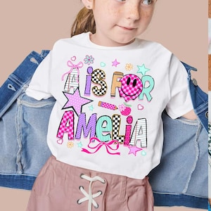 May include: A white t-shirt featuring a colorful graphic with the text A IS FOR AMELIA. The design includes a pink checkered smiley face, a pencil, stars, hearts, flowers, and bows in shades of pink, blue, orange, and black and white patterns.
