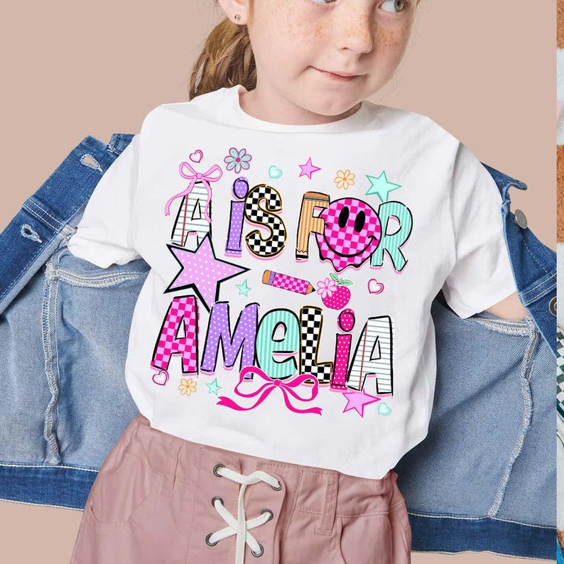 May include: A white t-shirt featuring a colorful graphic with the text A IS FOR AMELIA. The design includes a pink checkered smiley face, a pencil, stars, hearts, flowers, and bows in shades of pink, blue, orange, and black and white patterns.
