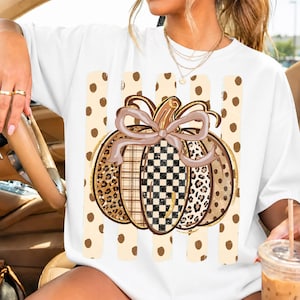 K&ouml;nnte beinhalten: Ein wei&szlig;es T-Shirt mit einer gro&szlig;en K&uuml;rbisgrafik und einer Schleife. Das K&uuml;rbisdesign enth&auml;lt Abschnitte mit Leopardenmuster, Karomuster sowie einem schwarz-wei&szlig;en Schachbrettmuster. Der Hintergrund der Grafik zeigt vertikale beige Punkte-Streifen.