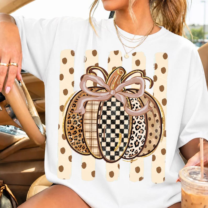 K&ouml;nnte beinhalten: Ein wei&szlig;es T-Shirt mit einer gro&szlig;en K&uuml;rbisgrafik und einer Schleife. Das K&uuml;rbisdesign enth&auml;lt Abschnitte mit Leopardenmuster, Karomuster sowie einem schwarz-wei&szlig;en Schachbrettmuster. Der Hintergrund der Grafik zeigt vertikale beige Punkte-Streifen.