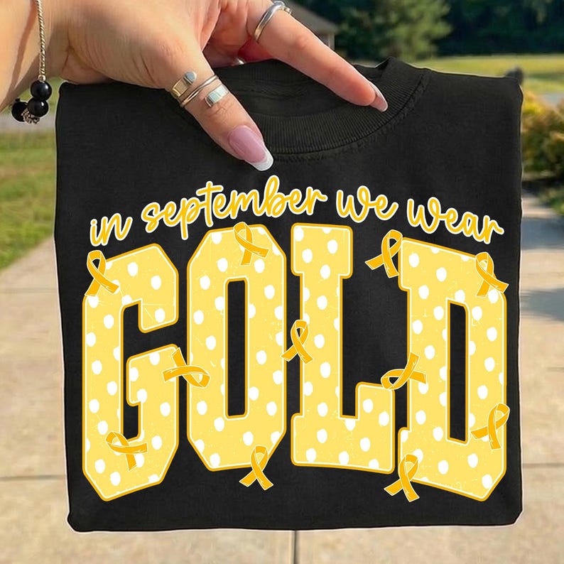 May include: A black t-shirt featuring the yellow text "in september we wear GOLD" in a bold, blocky font. The letters are filled with a white polka dot pattern and decorated with multiple small yellow awareness ribbons throughout the design.