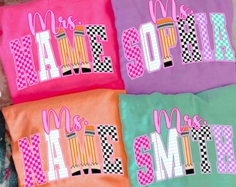 May include: Four brightly coloured t-shirts, each personalised for a teacher. The shirts display "Mrs." above a name, with a pencil graphic and chequered patterns. The colours are pink, purple, orange, and green.