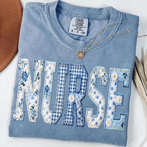 May include: A light blue Comfort Colors t-shirt with the word "NURSE" appliqued across the front. The letters are a mix of floral, polka dot, and gingham patterns in shades of blue and white. A gold necklace with a pendant rests on the shirt.
