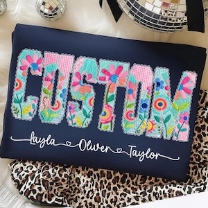 Op de afbeelding: Marineblauw kussen met het woord "CUSTOM" in kleurrijke bloemenpatchwork letters. Daaronder staan de namen "Layla", "Oliver" en "Taylor" in witte letters. Het kussen ligt op een luipaardprint stof.