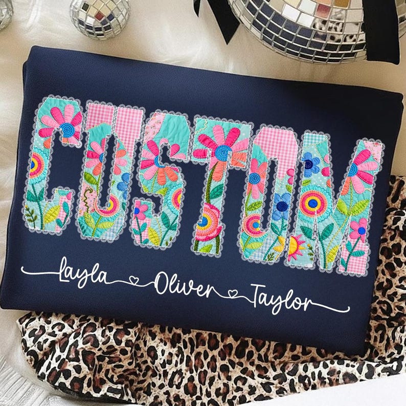 Op de afbeelding: Marineblauw kussen met het woord "CUSTOM" in kleurrijke bloemenpatchwork letters. Daaronder staan de namen "Layla", "Oliver" en "Taylor" in witte letters. Het kussen ligt op een luipaardprint stof.