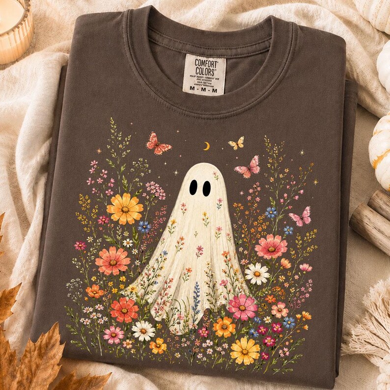 May include: A dark brown crewneck t-shirt featuring a central graphic of a white ghost surrounded by colorful wildflowers and butterflies. The shirt has a Comfort Colors brand label at the inner neckline and a short-sleeved, casual design.
