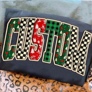 May include: A dark gray sweatshirt with the word "CUSTOM" in a festive design. The letters feature a mix of patterns, including green and red Christmas trees, black and white checkered squares, and red with white Christmas motifs.