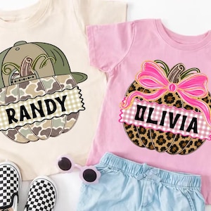 May include: A beige t-shirt featuring a camo pumpkin with a green baseball cap and the name RANDY, and a pink t-shirt featuring a leopard print pumpkin with a bright pink bow and the name OLIVIA, accompanied by checkered slip-on shoes and sunglasses.