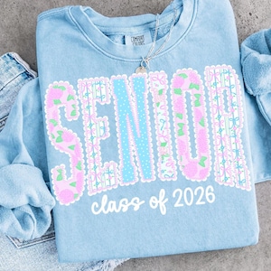 May include: Light blue sweatshirt with the word "SENIOR" in pink floral letters and a light blue dotted center. The text "class of 2026" is below the word "SENIOR". A silver necklace is on top of the sweatshirt.