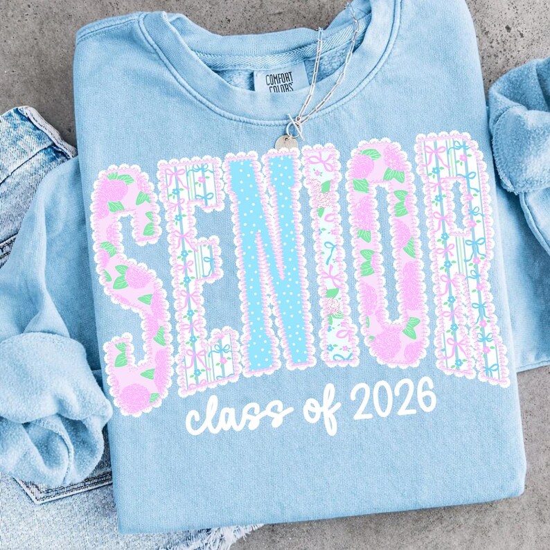 May include: Light blue sweatshirt with the word "SENIOR" in pink floral letters and a light blue dotted center. The text "class of 2026" is below the word "SENIOR". A silver necklace is on top of the sweatshirt.