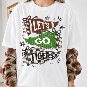 May include: A white short-sleeve t-shirt featuring a graphic of three pennants. The top brown pennant says LET'S, the middle green football field pennant says GO, and the bottom checkered pennant says TIGERS, accented with small stars.
