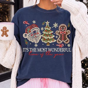 K&ouml;nnte beinhalten: Ein marineblaues Sweatshirt mit Rundhalsausschnitt und einem Aufdruck von Weihnachtsmann, geschm&uuml;cktem Weihnachtsbaum und Lebkuchenmann. Der Text lautet IT'S THE MOST WONDERFUL time of the year. Ein passender wei&szlig;er Becher ist ebenfalls zu sehen.