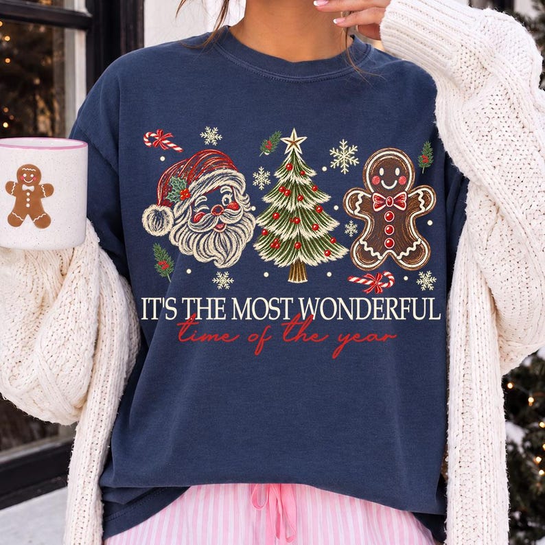 K&ouml;nnte beinhalten: Ein marineblaues Sweatshirt mit Rundhalsausschnitt und einem Aufdruck von Weihnachtsmann, geschm&uuml;cktem Weihnachtsbaum und Lebkuchenmann. Der Text lautet IT'S THE MOST WONDERFUL time of the year. Ein passender wei&szlig;er Becher ist ebenfalls zu sehen.