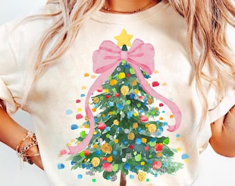 Könnte beinhalten: Ein weißes T-Shirt mit einer Aquarell-Illustration eines Weihnachtsbaums, der mit bunten Kugeln und einer großen rosa Schleife geschmückt ist. Der Baum hat einen gelben Stern oben.