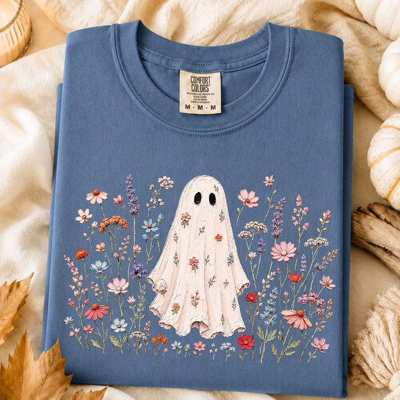 May include: A blue crew neck t-shirt with a Comfort Colors label at the inner collar. The front features a graphic of a white ghost surrounded by a variety of colorful wildflowers in shades of pink, orange, blue, and purple, with small floral patterns on the ghost.