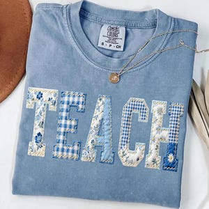 Op de afbeelding: Een lichtblauw Comfort Colors T-shirt met het woord "TEACH" in een patchwork-ontwerp. De letters hebben bloemen- en ruitpatronen in blauw en wit. Een gouden ketting met een hanger ligt op het shirt.