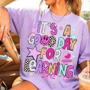 May include: A light purple t-shirt featuring the text IT'S A GOOD DAY FOR LEARNING in a multi-colored, patterned font. The design includes graphic elements like a smiley face, stars, bows, a pencil, and a strawberry, all in shades of pink, blue, and tan.