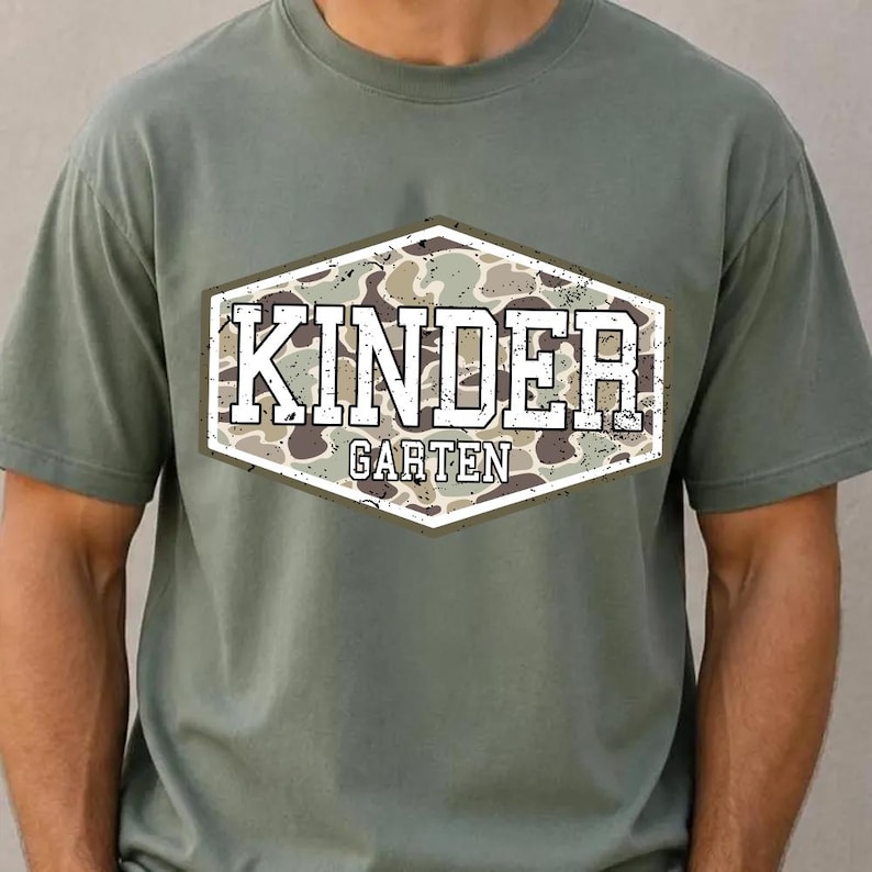 May include: Olive green t-shirt with a camouflage design in a white-bordered, hexagonal shape. The text "KINDER GARTEN" is printed in white block letters above the word "GARTEN". The shirt has short sleeves and a crew neck.