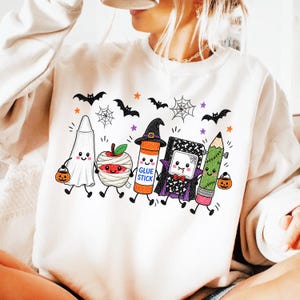 Pode incluir: Uma camisola cor de creme com um gr&aacute;fico de cinco artigos escolares antropom&oacute;rficos vestidos para o Halloween: um fantasma, uma ma&ccedil;&atilde; m&uacute;mia, uma cola em barra com chap&eacute;u de bruxa, um caderno vampiro e um l&aacute;pis monstro, rodeados de morcegos.