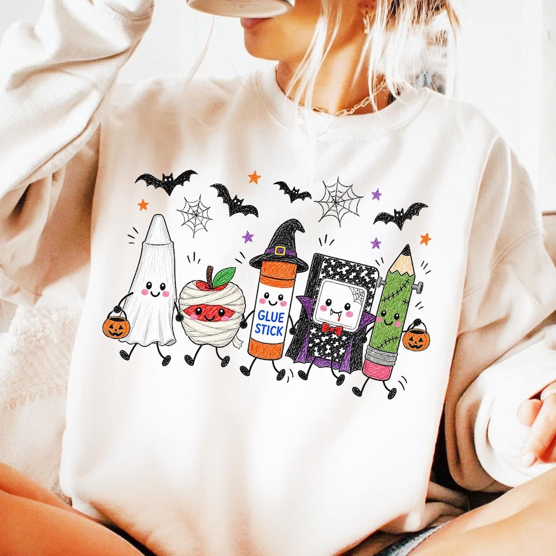Pode incluir: Uma camisola cor de creme com um gr&aacute;fico de cinco artigos escolares antropom&oacute;rficos vestidos para o Halloween: um fantasma, uma ma&ccedil;&atilde; m&uacute;mia, uma cola em barra com chap&eacute;u de bruxa, um caderno vampiro e um l&aacute;pis monstro, rodeados de morcegos.